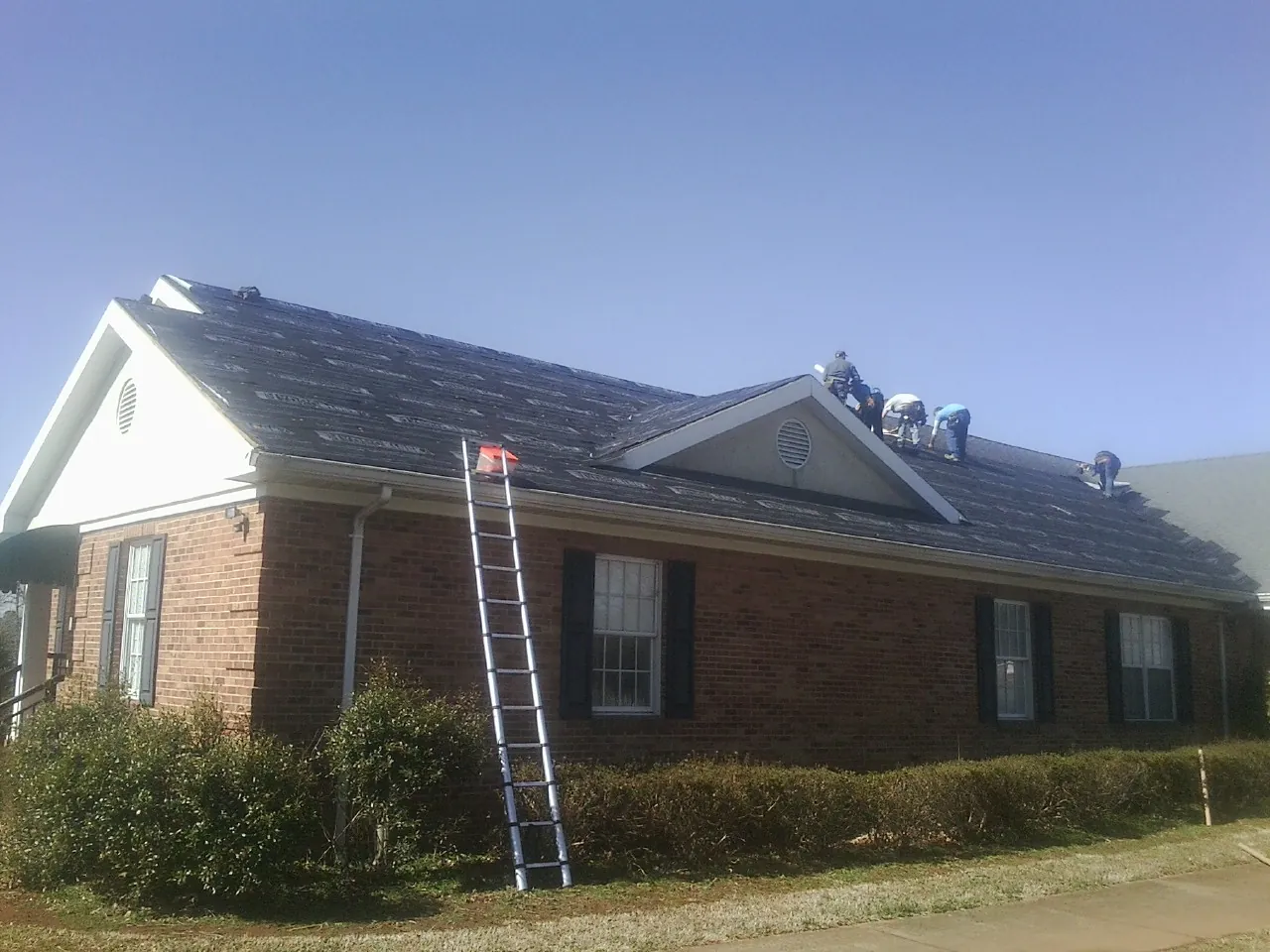 Crew tearing shingles off a brick ranch roof with a ladder set — Emergency Roof Repair work by our crew in Rowe