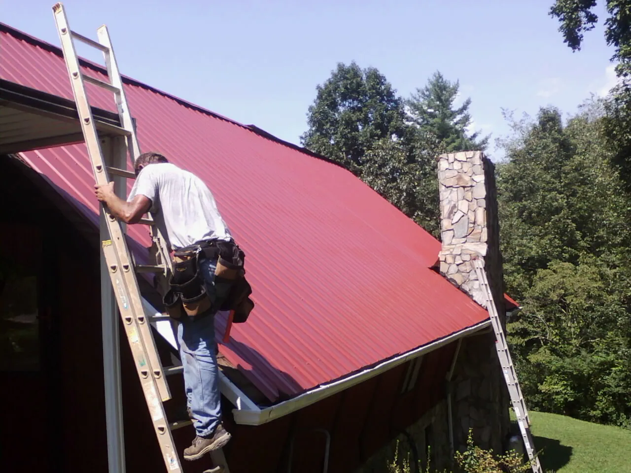 Roofer on a ladder beside a red metal roof and stone chimney — Soffit & Fascia Repair by Roofing Brothers in Rowe
